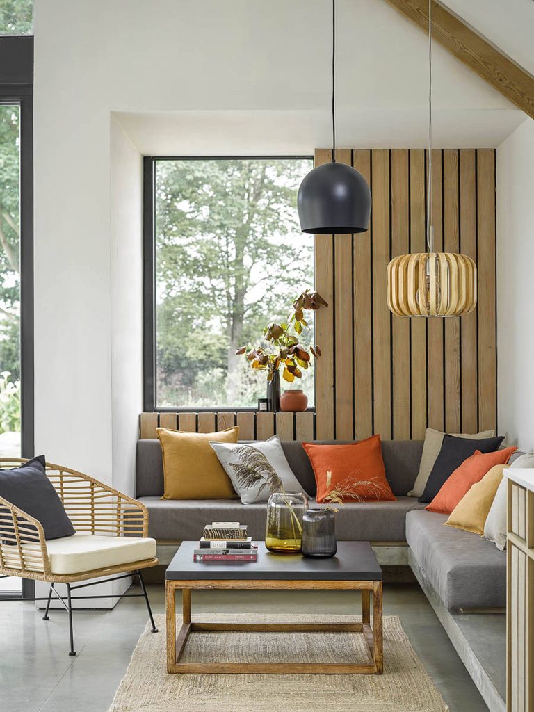 A corner sofa with cushions in yellow and orange. Two pendants hang above, with wooden panelling around a window in the background
